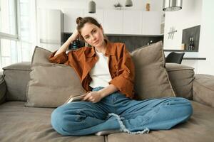 portret van jong modern vrouw, vrouw leerling zittend Bij huis Aan sofa en op zoek Bij camera, poseren Aan bank, glimlachen foto