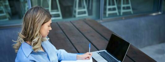 portret van zakenvrouw nemen notities, aan het studeren Aan haar computer, zittend buiten in park. jong vrouw schrijven omlaag, video conferenties Aan laptop foto