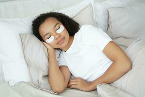 zwart vrouw met oog patches rust in bed, genieten van een schoonheid routine- foto