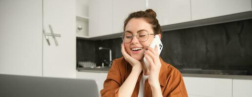 portret van mooi jong vrouw beheren haar eigen bedrijf van huis, freelancer maken telefoon telefoongesprek, zittend in keuken met laptop en glimlachen foto