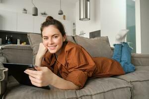 portret van schattig jong vrouw ontspannende Bij huis, aan het kijken film Aan digitaal tablet of lezing e-boek, gebruik makend van apparaatje terwijl aan het liegen Aan bankstel in leven kamer, glimlachen Bij camera foto