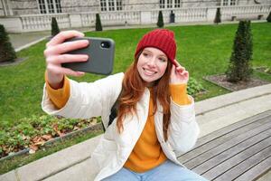 jong tiener roodharige meisje zit Aan bank in park en duurt selfie, maakt een foto van haarzelf met smartphone app, records vlog