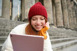 portret van jong roodharige vrouw zittend buitenshuis Aan trap, lezing ebook Aan digitaal tablet, vervelend rood hoed en warm kleren foto