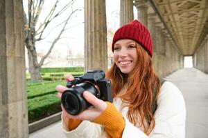 jong roodharige vrouw fotograaf, maakt levensstijl het schieten in stad centrum, duurt foto's en glimlacht, looks voor perfect schot, maakt afbeelding foto