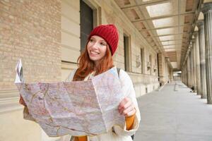 glimlachen jong roodharige vrouw in rood hoed, looks Bij papier kaart naar kijken voor toerist attractie. toerisme en mensen concept. meisje onderzoekt stad, geprobeerd naar vind manier foto