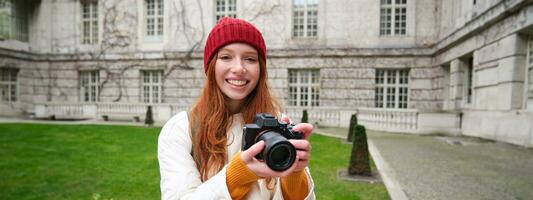 roodharige meisje fotograaf duurt foto's Aan professioneel camera buitenshuis, vangt Street Style schoten, looks opgewonden terwijl nemen afbeeldingen foto