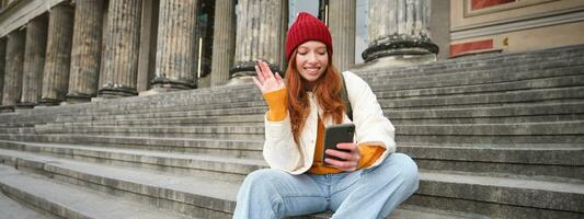 roodharige vrouw toerist praat Aan mobiel telefoon, video chatten met vrienden, zegt Hoi en golven hand- Bij smartphone camera foto