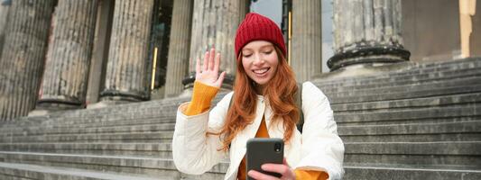 jong roodharige vrouw zit Aan trap buitenshuis en golven hand- Bij smartphone camera, video chats met vrienden, verbindt naar openbaar Wifi foto