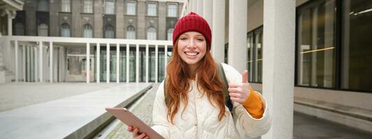 gelukkig roodharige meisje in rood hoed, wandelingen in de omgeving van stad met digitaal tablet, verbindt naar openbaar internet Wifi en looks voor route, looks Bij kaart Aan haar apparaatje foto