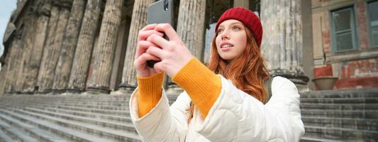 portret van jong roodharige vrouw duurt foto's Aan haar mobiel telefoon camera, gebruik makend van smartphone naar vastleggen mooi zo schot buitenshuis foto