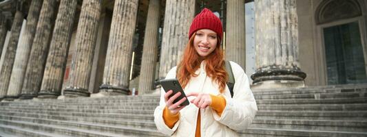 portret van jong roodharige vrouw Holding mobiel telefoon, gebruik makend van smartphone app, wandelen in stad centrum, gebruik makend van kaart toepassing of sms'en een bericht foto