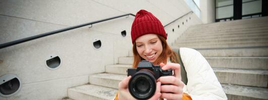 stedelijk mensen en levensstijl. gelukkig roodharige vrouw duurt foto's, Holding professioneel digitaal camera, fotograferen Aan straten foto