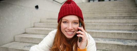 mooi glimlachen roodharige vrouw model, zit Aan straat en praat Aan mobiel telefoon, toepassingen smartphone app naar telefoontje Buitenland, lachend gedurende telefoon gesprek foto
