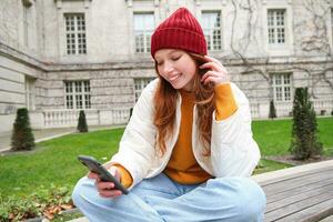 jong glimlachen roodharige meisje zit Aan bank en toepassingen smartphone app, leest nieuws online, horloges video Aan mobiel telefoon terwijl ontspannen in park foto