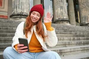 roodharige vrouw toerist praat Aan mobiel telefoon, video chatten met vrienden, zegt Hoi en golven hand- Bij smartphone camera foto