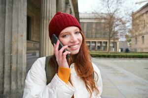 glimlachen jong roodharige vrouw luistert naar stem bericht, maakt een telefoon telefoongesprek, wandelingen Aan straat en praat naar iemand Aan smartphone foto
