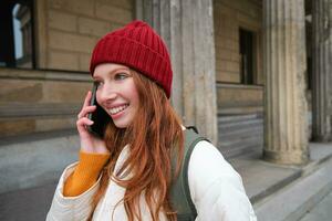 glimlachen jong roodharige vrouw luistert naar stem bericht, maakt een telefoon telefoongesprek, wandelingen Aan straat en praat naar iemand Aan smartphone foto