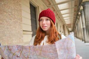 avontuurlijk roodharige meisje wandelingen in stad- met papier kaart, onderzoekt stad net zo toerist, looks voor populair toerisme attracties, looks in de omgeving van opgewonden en glimlacht foto