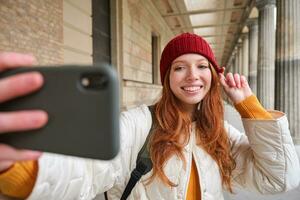 schattig jong roodharige vrouw duurt selfie Aan straat met mobiel telefoon, maakt een foto van haarzelf met smartphone app Aan straat