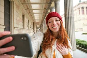 schattig jong roodharige vrouw duurt selfie Aan straat met mobiel telefoon, maakt een foto van haarzelf met smartphone app Aan straat
