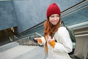 portret van roodharige meisje verkennen stad met smartphone kaart app. jong vrouw gebruik makend van mobiel telefoon en wandelen in de omgeving van stad, gaan naar beneden trap foto