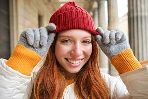 hoofdschot van gelukkig roodharige meisje met sproeten, draagt rood hoed en handschoenen in winter, wandelingen in de omgeving van stad Aan koud weer en glimlacht foto