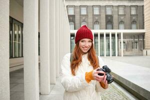 glimlachen roodharige meisje fotograaf, nemen afbeeldingen in stad, maakt foto's buitenshuis Aan professioneel camera foto