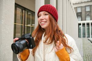 glimlachen roodharige meisje fotograaf, nemen afbeeldingen in stad, maakt foto's buitenshuis Aan professioneel camera foto