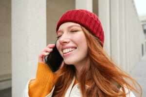 mobiel breedband en mensen. glimlachen jong roodharige vrouw wandelingen in stad- en praat Aan mobiel telefoon, roeping vriend Aan smartphone, gebruik makend van internet naar maken een telefoontje Buitenland foto