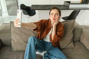 portret van schattig glimlachen vrouw model, duurt selfie Aan smartphone app, posten foto's Aan sociaal media, gebruik makend van mobiel telefoon camera, poseren Aan sofa Bij huis foto