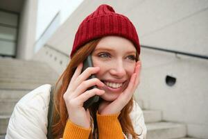 jong elegant roodharige meisje in rood hoed, zit Aan straat en praat Aan mobiel telefoon, heeft telefoon gesprek, ringen haar vriend terwijl ontspant buitenshuis foto