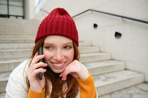 mooi glimlachen roodharige vrouw model, zit Aan straat en praat Aan mobiel telefoon, toepassingen smartphone app naar telefoontje Buitenland, lachend gedurende telefoon gesprek foto