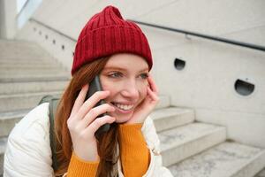 jong elegant roodharige meisje in rood hoed, zit Aan straat en praat Aan mobiel telefoon, heeft telefoon gesprek, ringen haar vriend terwijl ontspant buitenshuis foto