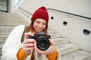 stedelijk mensen en levensstijl. gelukkig roodharige vrouw duurt foto's, Holding professioneel digitaal camera, fotograferen Aan straten foto