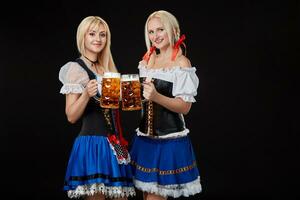 twee mooi blond Dames zijn Holding bril van bier in handen en staan Aan zwart achtergrond in studio. foto