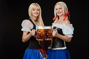 twee mooi blond Dames zijn Holding bril van bier in handen en staan Aan zwart achtergrond in studio. foto
