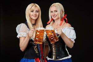 twee mooi blond Dames zijn Holding bril van bier in handen en staan Aan zwart achtergrond in studio. foto