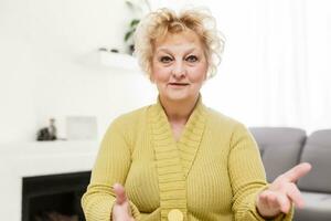 glimlachen midden- oud volwassen grijs haren vrouw op zoek Bij camera, gelukkig oud dame poseren Bij huis binnen, positief single senior gepensioneerd vrouw zittend Aan sofa in leven kamer hoofdschot portret foto