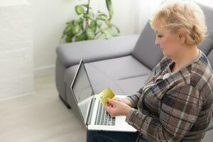 oud vrouw Holding een credit kaart in voorkant van laptop met blanco scherm foto