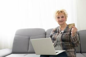 oud vrouw Holding een credit kaart in voorkant van laptop met blanco scherm foto