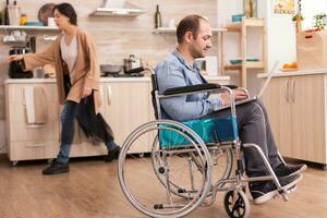 ongeldig Mens in rolstoel werken van een afstand Aan laptop in keuken terwijl is staand in voorkant van koelkast. gehandicapt verlamd gehandicapten Mens met wandelen onbekwaamheid integreren na een ongeluk. foto