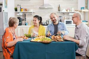 moeder en dochter glimlachen terwijl hebben heerlijk lunch in keuken met familie. senior Mens Holding wijn glas. smakelijk geroosterd aardappelen. foto