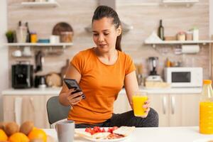 vrouw drinken vers oranje sap in de ochtend- terwijl browsen Aan smartphone in keuken. huisvrouw gebruik makend van modern technologie en drinken gezond, natuurlijk, eigengemaakt oranje sap. verfrissend ochtend- foto