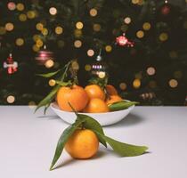 vers mandarijnen in een bord Aan een wit tafel achter Daar is een Kerstmis boom met lichten en speelgoed. Kerstmis achtergrond. foto