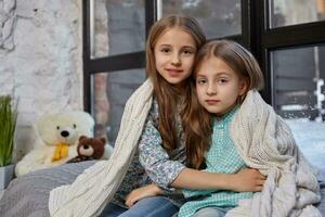 de beeld van twee schattig weinig zussen zittend Aan vensterbank, gedekt door deken, knuffelen elk andere foto