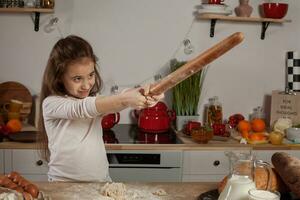 mooi weinig meisje gekleed in een wit blouse is maken een deeg voor bakken een brood Bij een keuken. foto