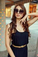 detailopname portret van jong elegant brunette in zwart jurk en zonnebril, aanraken haar gezicht. mode straat schot foto
