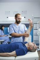 mannetje ouderen geduldig ontvangen osteopathisch zorg van een medisch beoefenaar voor arm pijn. Kaukasisch gepensioneerd oud Mens geholpen door verpleegster in blauw scrubs door fysiotherapie oefening. foto