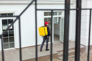 levering Mens staand met geel thermo rugzak voor voedsel levering in de buurt de Ingang huis met leeg ruimte naar kopiëren Plakken foto