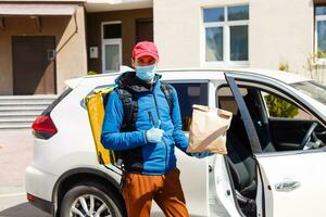 voedsel levering Mens in een beschermend masker en handschoenen met een thermo rugzak in de buurt een auto gedurende de quarantaine periode foto